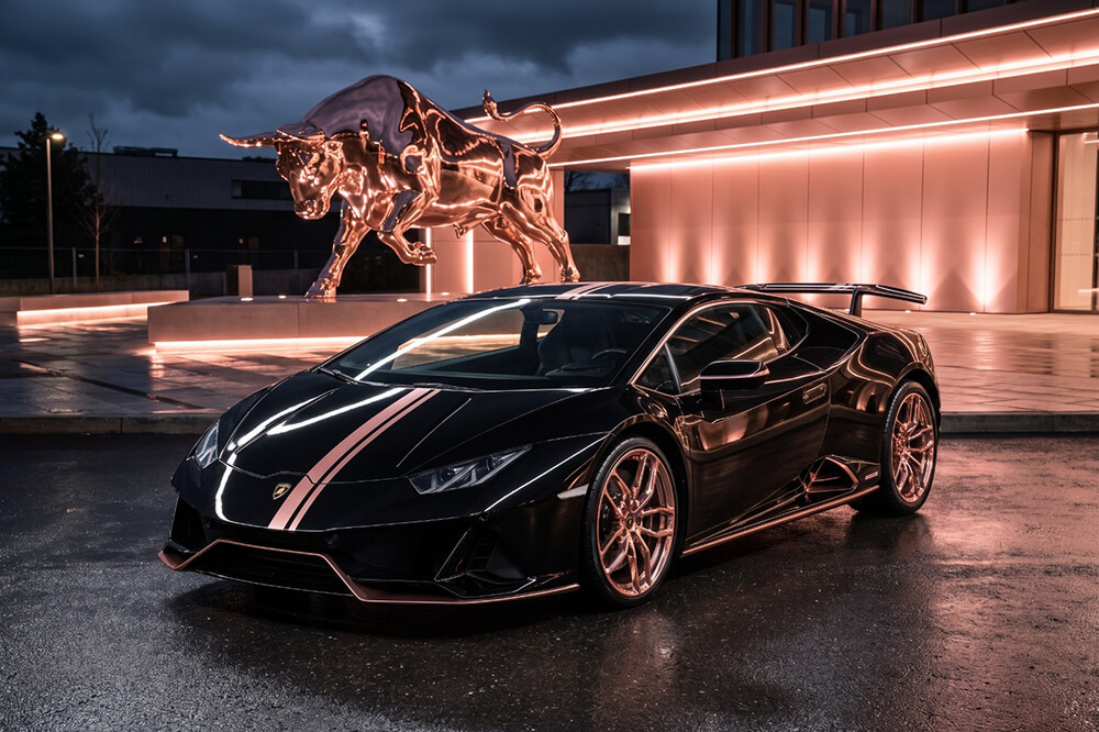Black Lamborghini with white racing stripe parked on a wet street near a glowing bull sculpture at dusk/dawn light.
