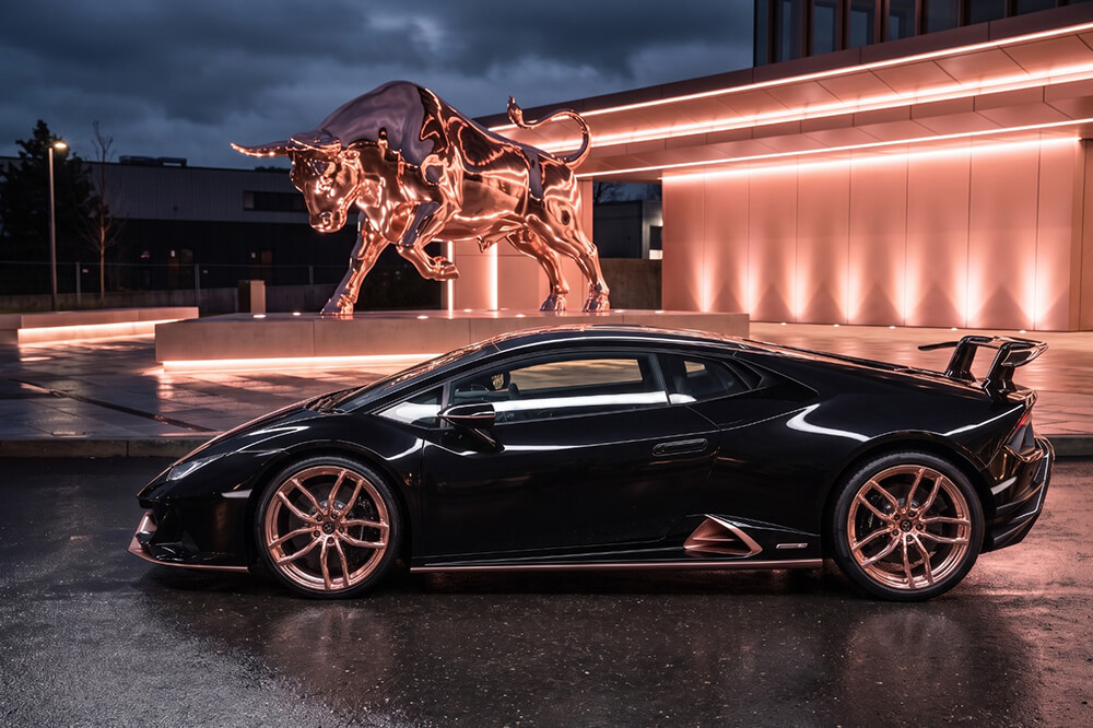 Black sports car parked on a wet street at night, illuminated by neon lights and a glowing bull sculpture in the background