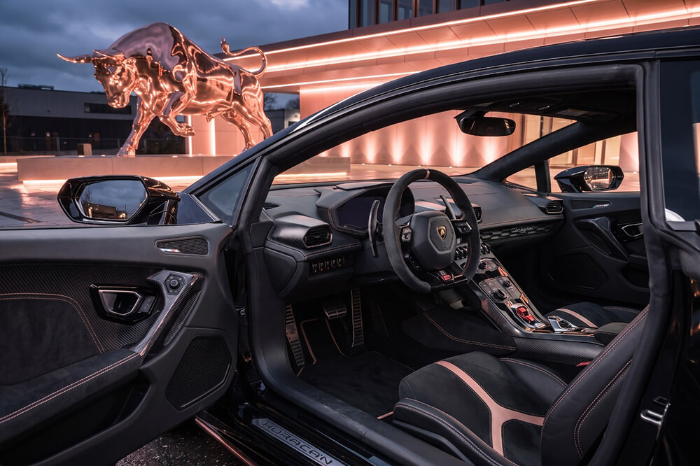 Lamborghini interior with black leather seats and red contrast stitching, open door, neon-lit showroom exterior with a chrome bull statue outside.